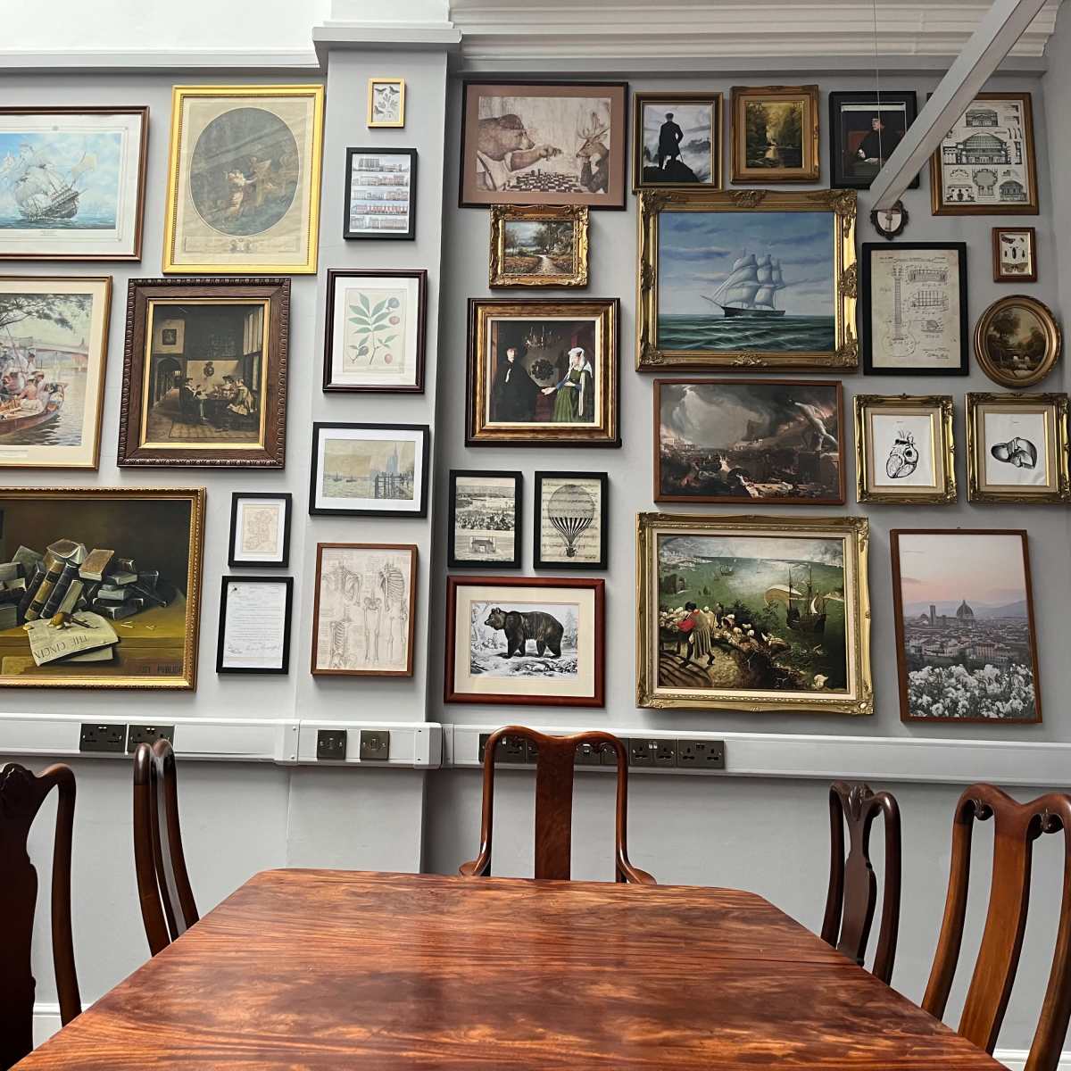 A dining area with a wooden table surrounded by chairs, set against a wall covered with a variety of framed artworks and photographs.