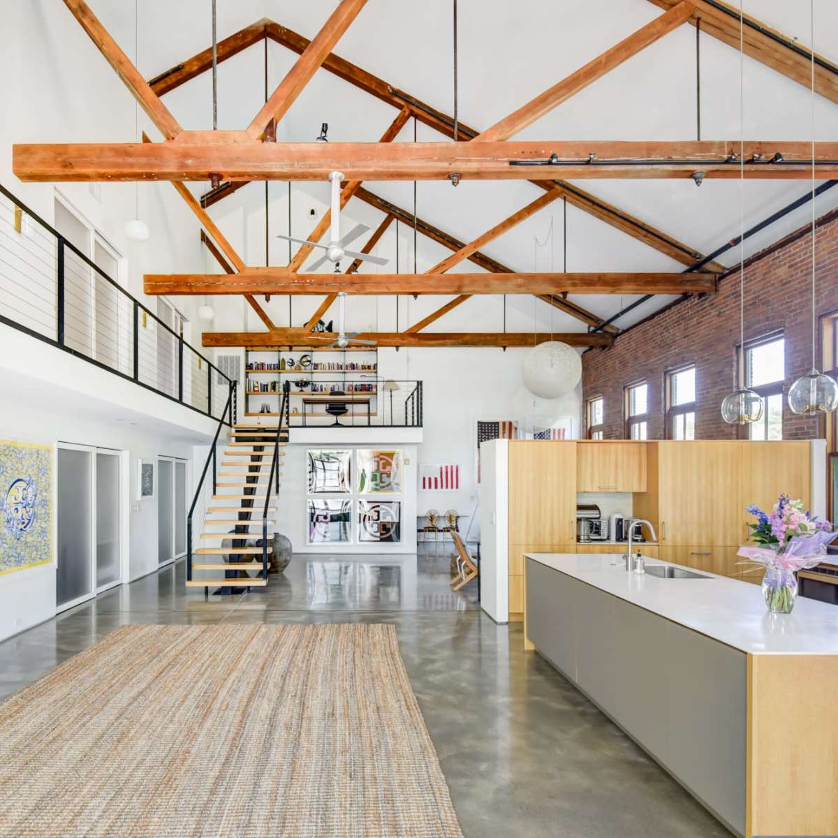 A spacious industrial-style loft featuring high wooden beams, an open kitchen with a large island, and a staircase leading to an upper level.