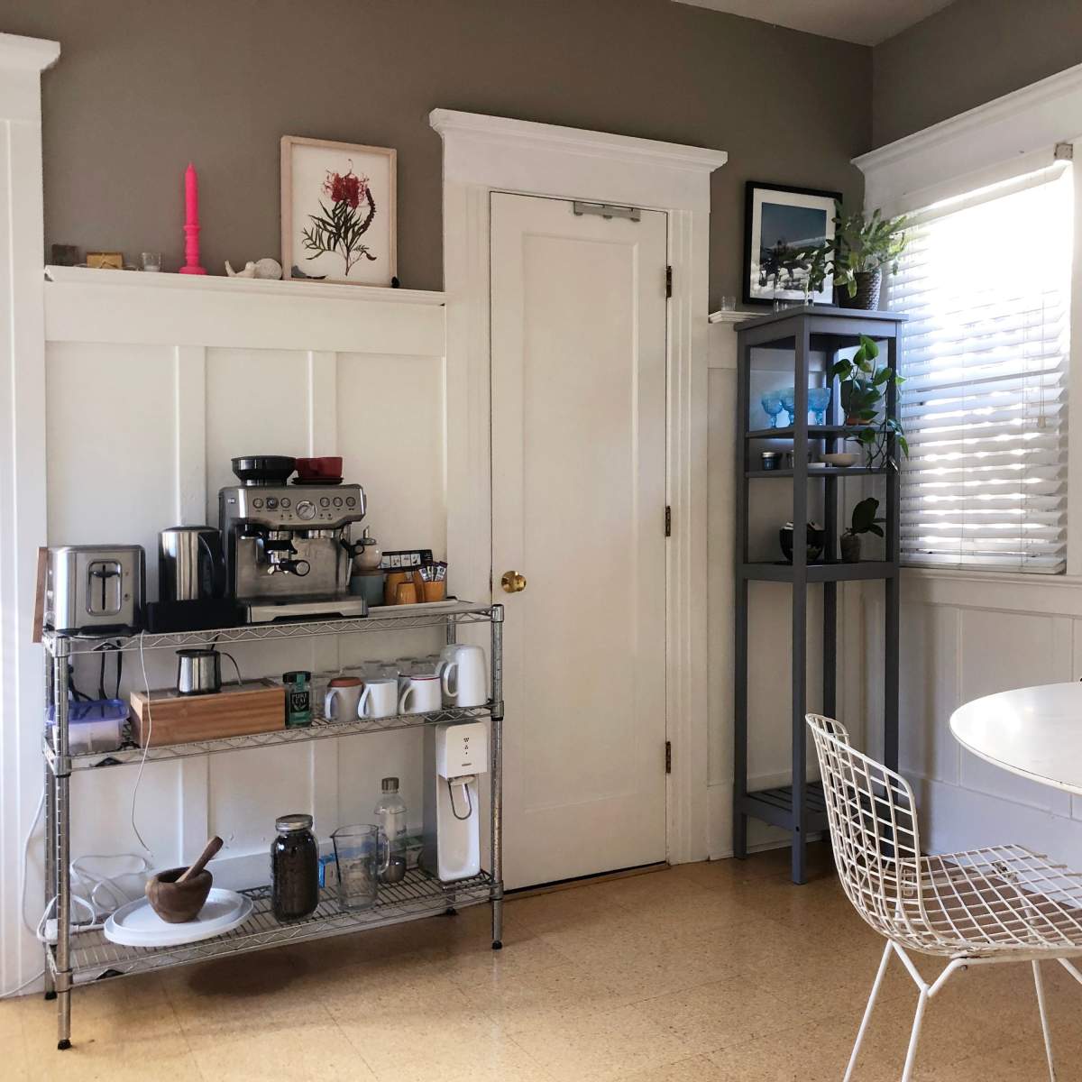 A kitchen area featuring a coffee station, a table with white chairs, and shelves holding plants and decorative items.