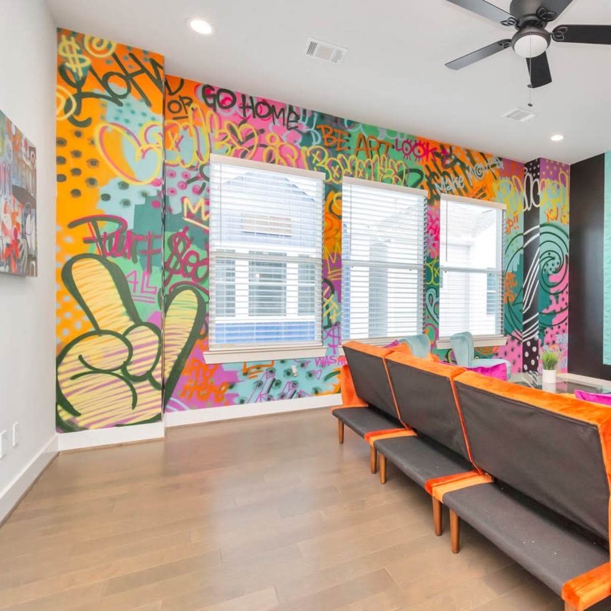 The room features a modern design with a colorful graffiti-style mural on one wall, large windows, and a black and orange sectional sofa facing a mounted television.