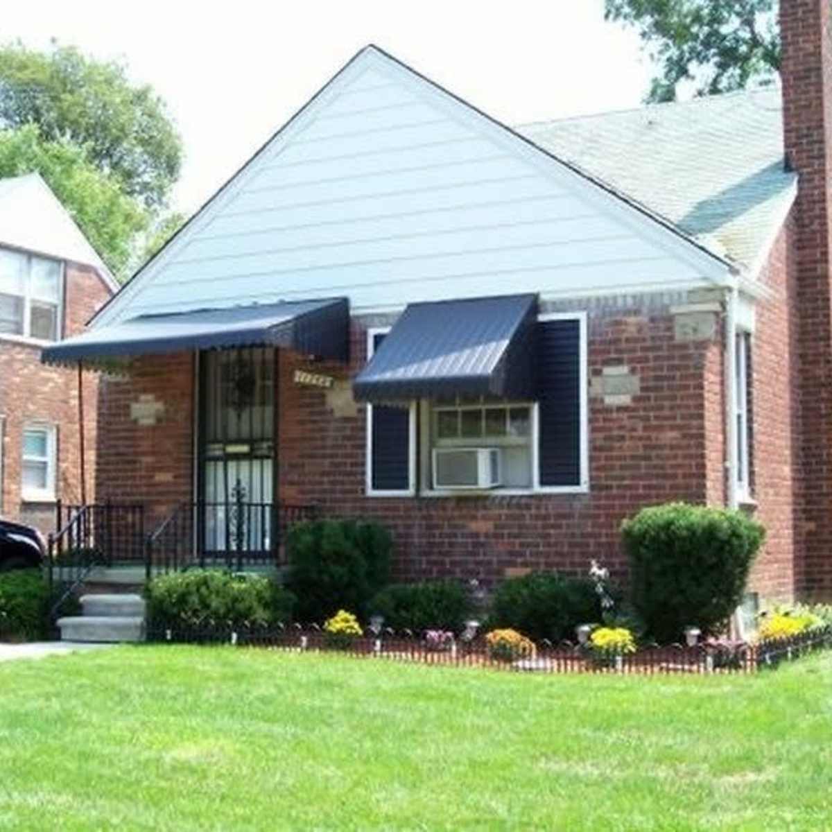 A brick house with white siding, a black awning over the front door, and well-maintained landscaping featuring colorful flowers and a green lawn.