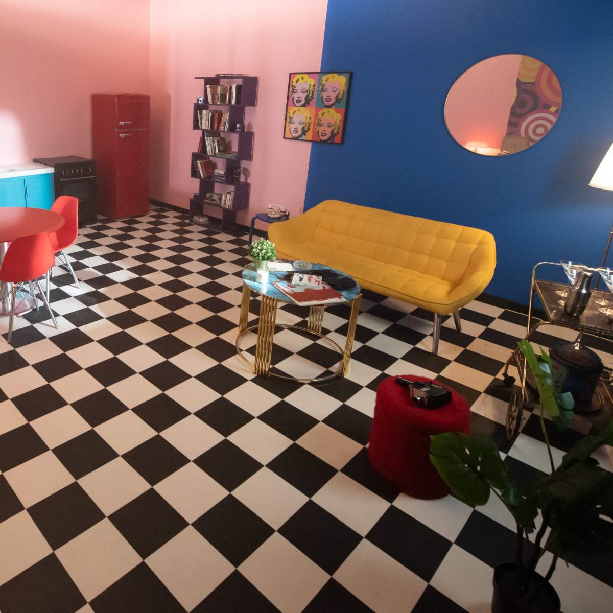 A brightly colored room with a checkered black-and-white floor featuring a blue accent wall, a yellow couch, red dining chairs, and a small kitchenette.