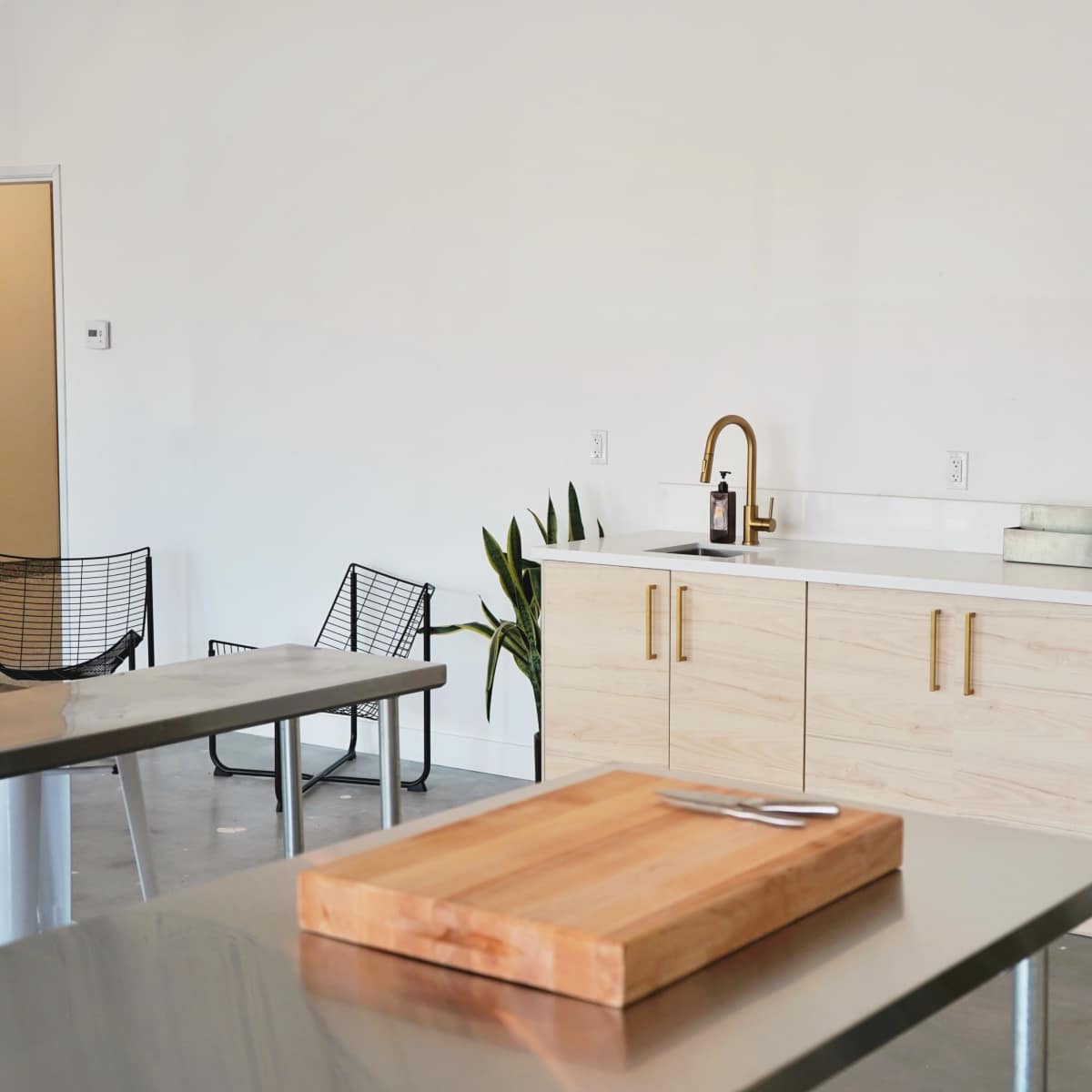 A modern kitchen with a wooden cabinet and two sinks is paired with a cutting board on a stainless steel table and black wire chairs nearby.