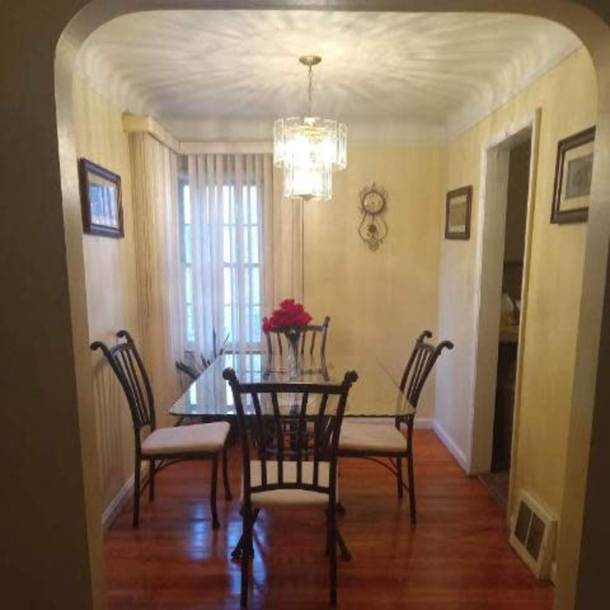 A dining area with a glass table surrounded by six chairs, illuminated by a chandelier, and a floral centerpiece on the table.