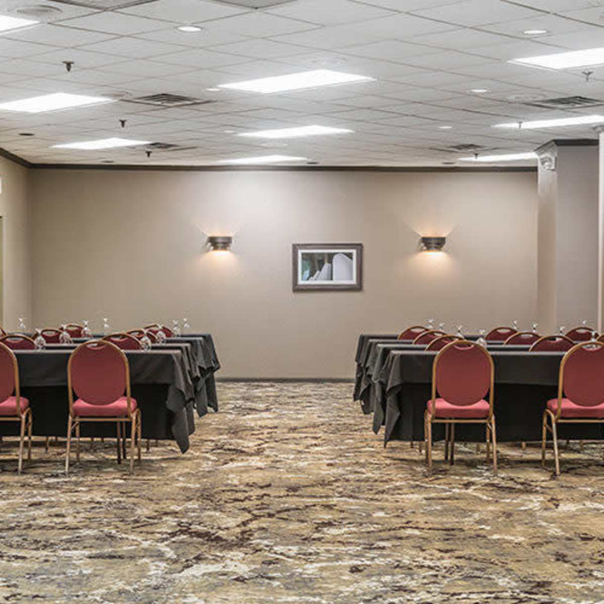 A meeting room features two rows of tables with red chairs and neutral-colored walls, illuminated by overhead lights.