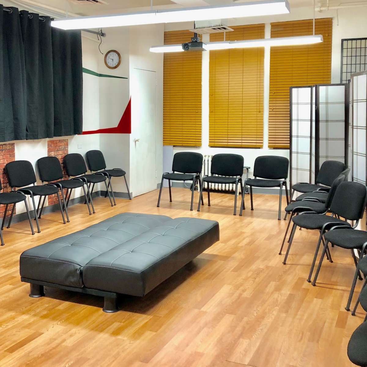 A room arranged with chairs in a circular formation around a rectangular black couch, with wooden flooring and partially closed blinds on the windows.