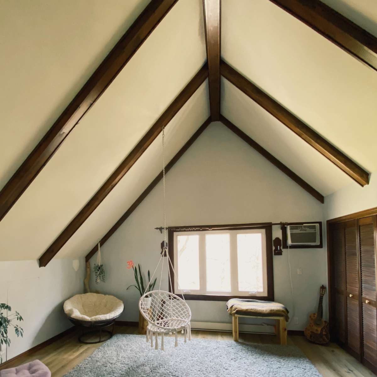The room features a high, sloped ceiling with wooden beams, a hanging chair, and a large window allowing natural light to enter.