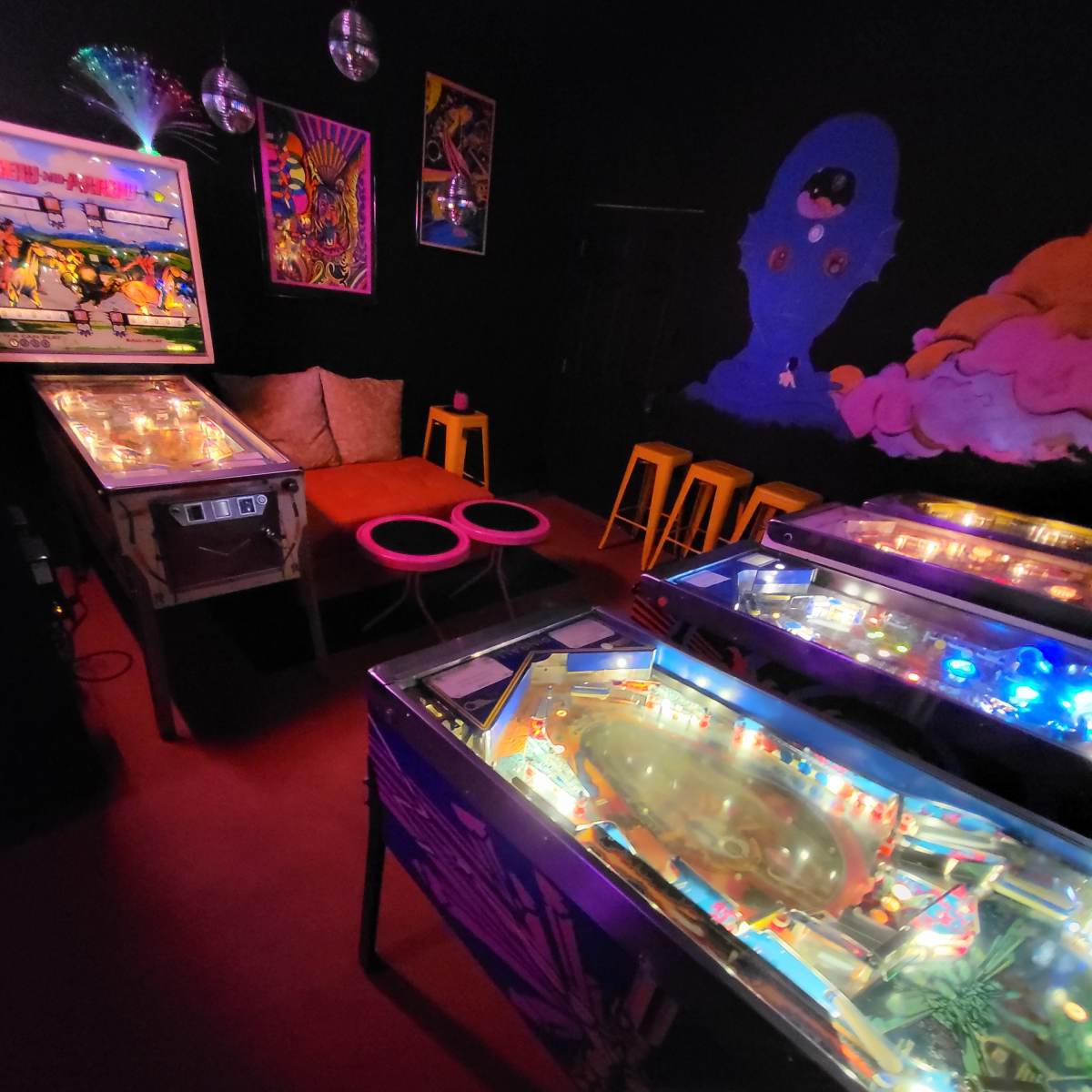 A dimly lit room filled with illuminated pinball machines, colorful artwork on the walls, and seating arrangements in the background.