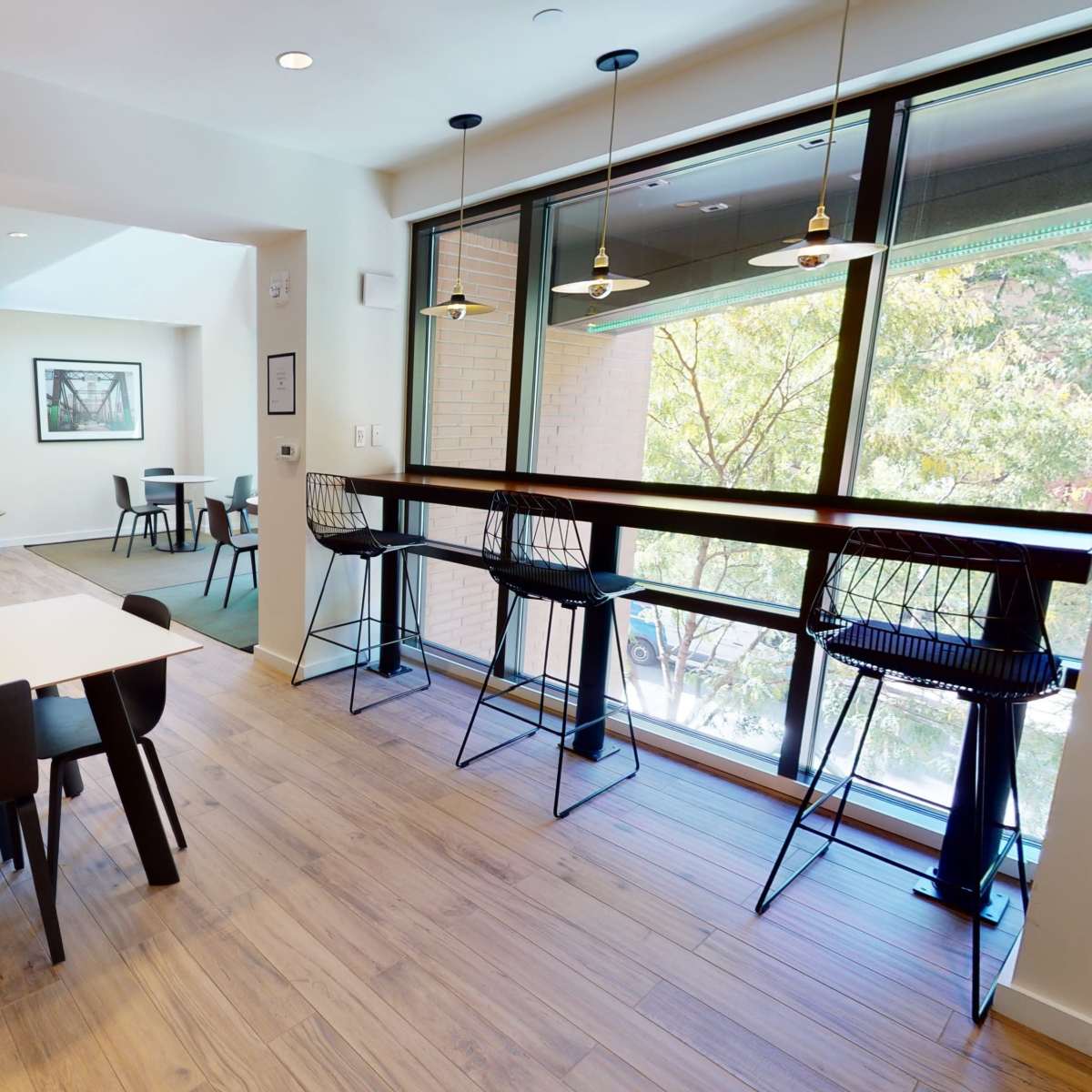 A modern, well-lit workspace features tables with chairs, a seating area with a sofa, and large windows overlooking greenery.
