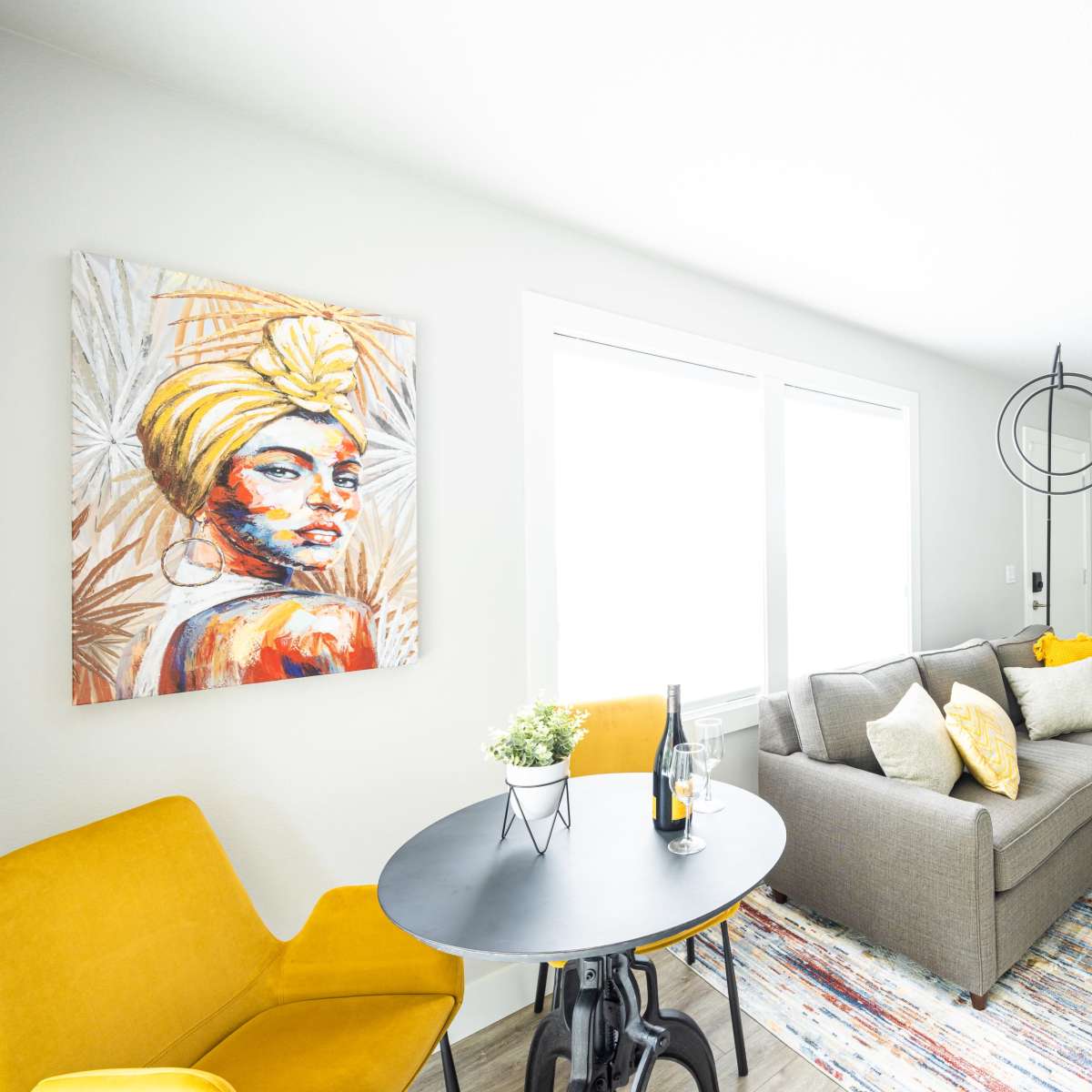A modern living room with a grey couch, yellow accent chairs, a circular black table, and colorful artwork featuring a woman on the wall.
