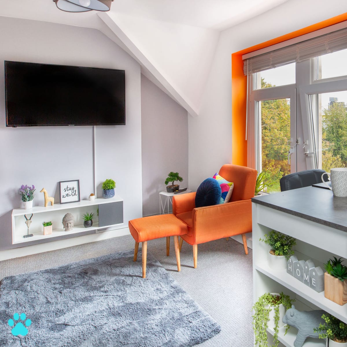 A modern living space features an orange-accented chair, a gray rug, a small dining table set with fruit, and large windows showcasing greenery outside.