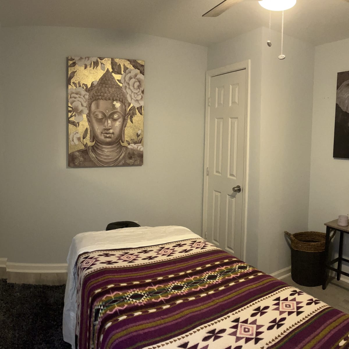A serene room with a massage table covered by a patterned blanket, a Buddha artwork on the wall, and an elephant picture beside a small plant and a woven basket.