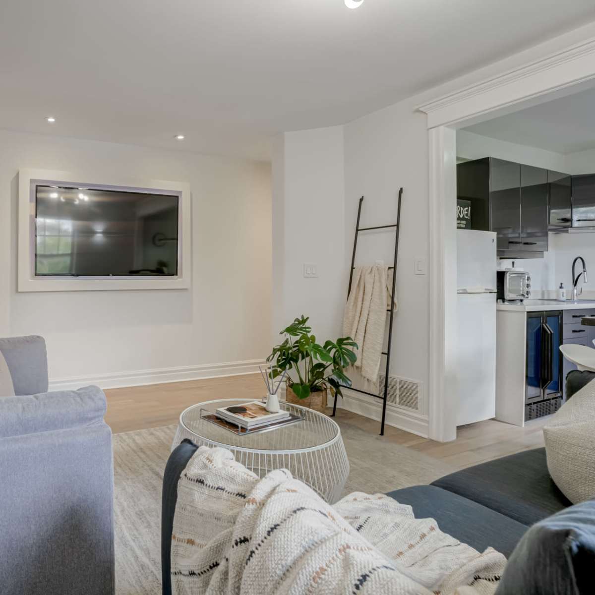 A modern living room features a gray sofa, a round coffee table, and a wall-mounted TV, with an open view toward a kitchen area.