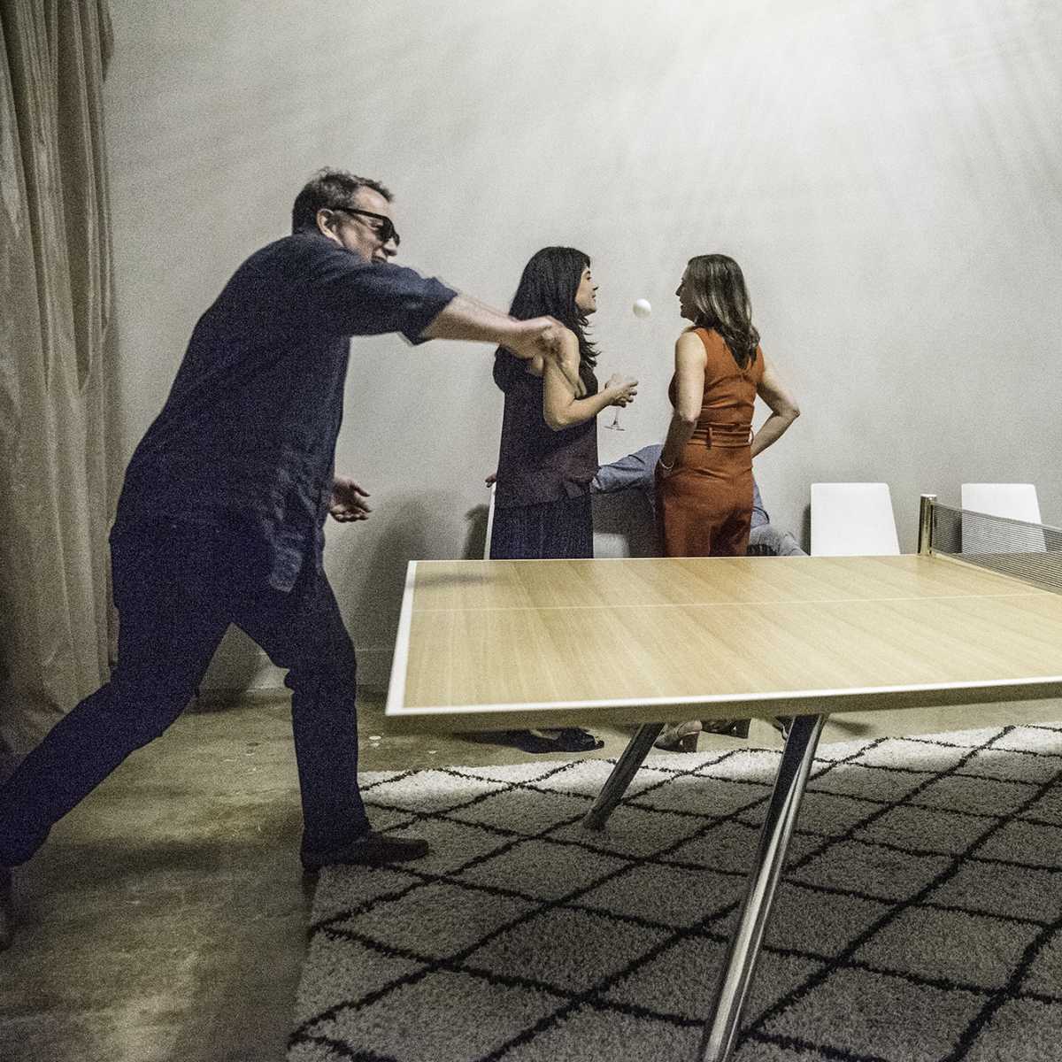 Four people are playing ping pong around a large table in a well-lit room with a rug on the floor.