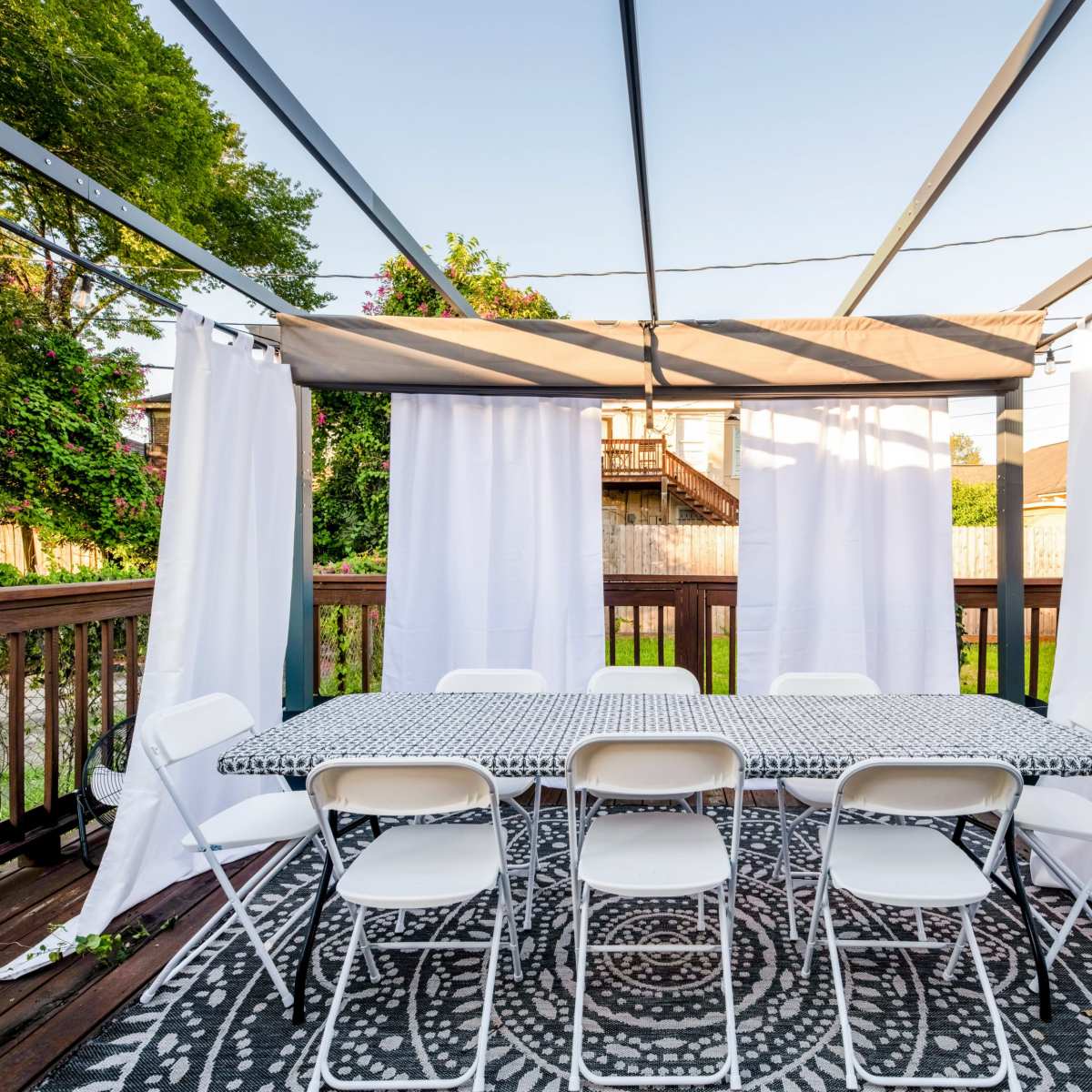 A large outdoor dining area is set with a patterned tablecloth and surrounded by white curtains and folding chairs on a wooden deck.