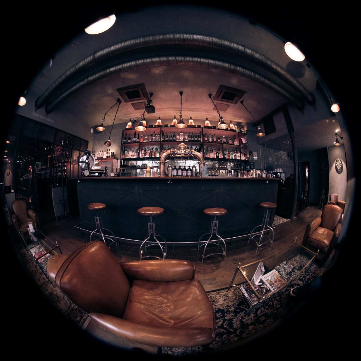 A vintage-style bar with a long counter, high stools, and shelves filled with bottles is depicted in a cozy interior setting.