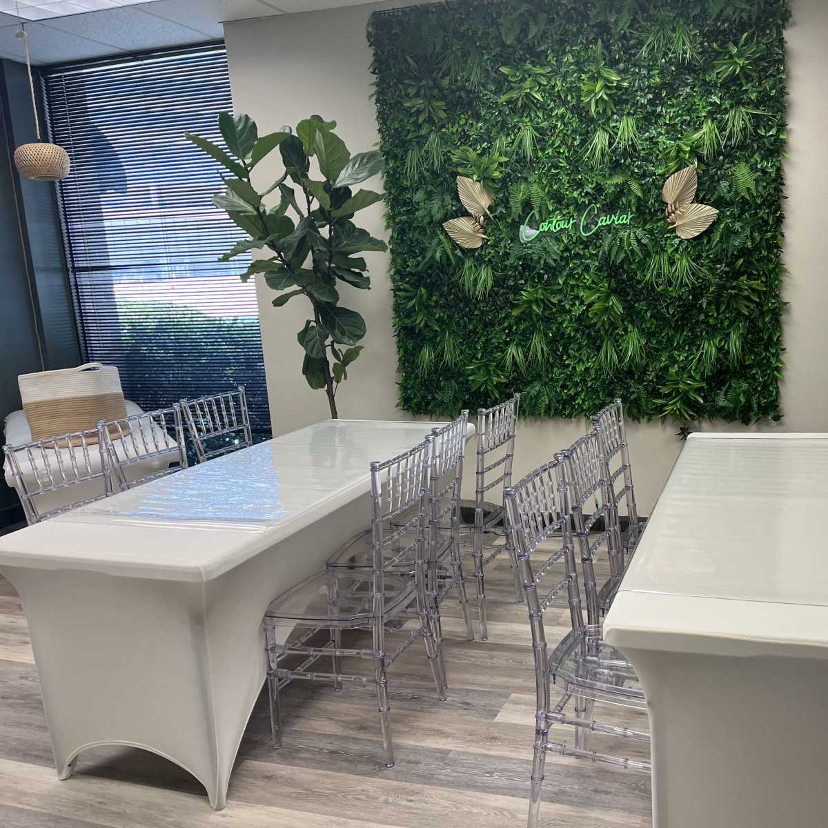 A modern room with two white tables, clear plastic chairs, and a green wall adorned with plants and decorative elements.