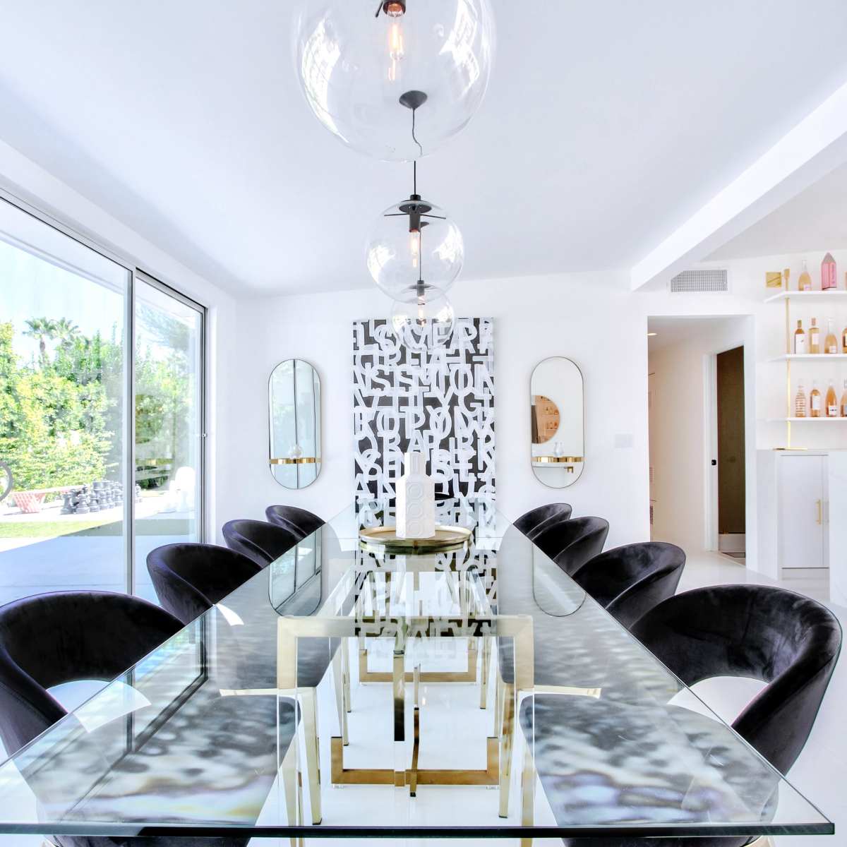 A modern dining room features a glass table surrounded by black chairs, with large windows overlooking a patio and greenery.