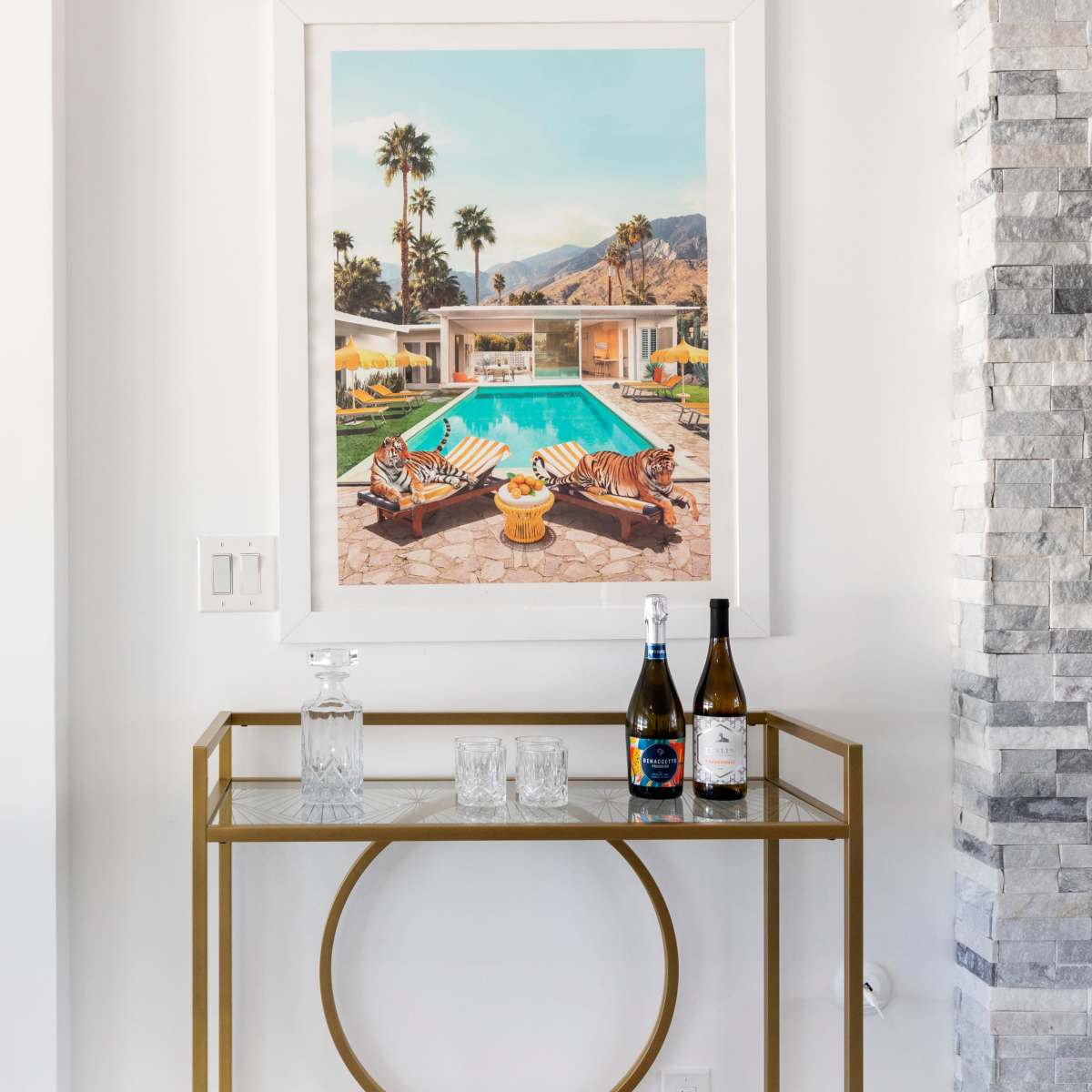 A stylish bar cart with glassware, bottles of wine, and an art piece of a poolside scene is placed against a white wall with stone accents.