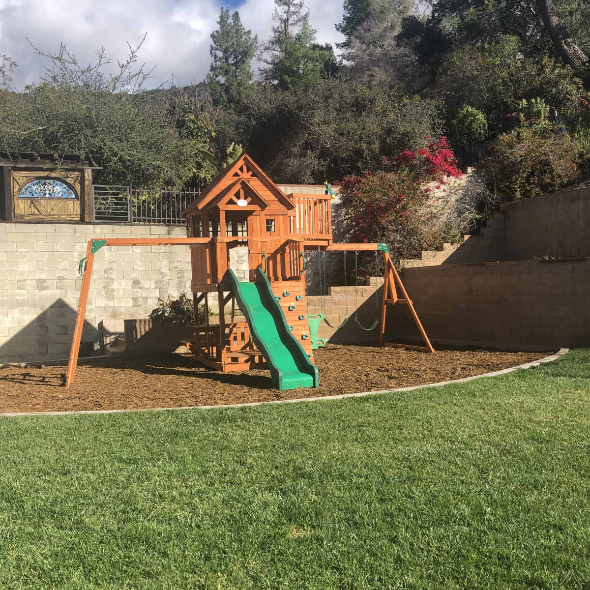 A wooden playground structure with a slide and swings is situated on a grassy area surrounded by a stone wall and greenery.