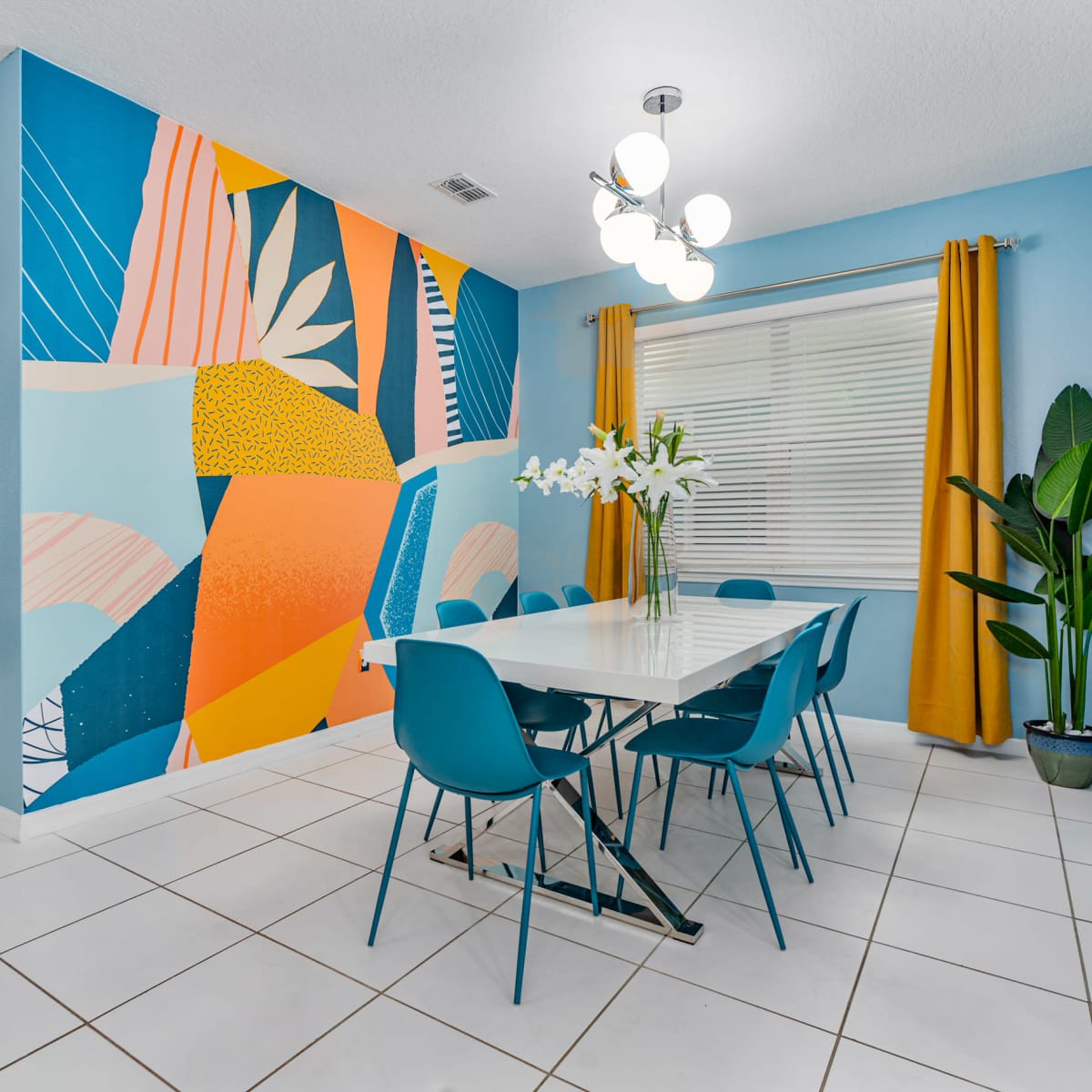 A modern dining area features a large white table surrounded by blue chairs, against a vibrant mural and bright yellow curtains.