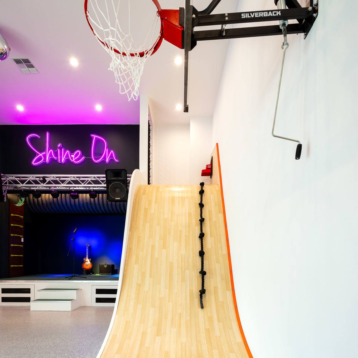 A basketball hoop next to a smooth wooden ramp with a black grip for climbing, set against a brightly lit wall with neon text.