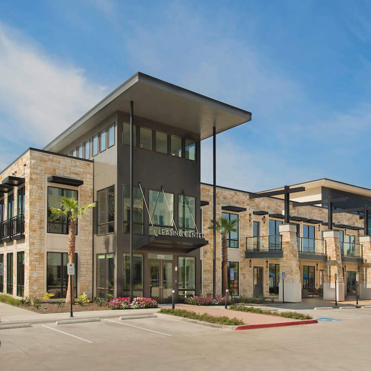 A modern two-story commercial building features large glass windows, a stone facade, and palm trees in the surrounding landscaping.