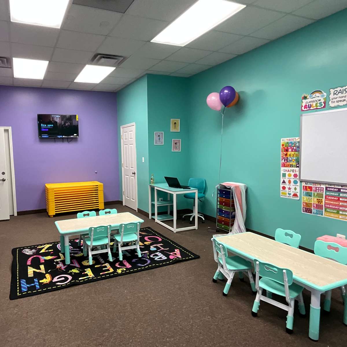 A colorful classroom features small tables and chairs, a play kitchen, storage units, and a television on the wall.