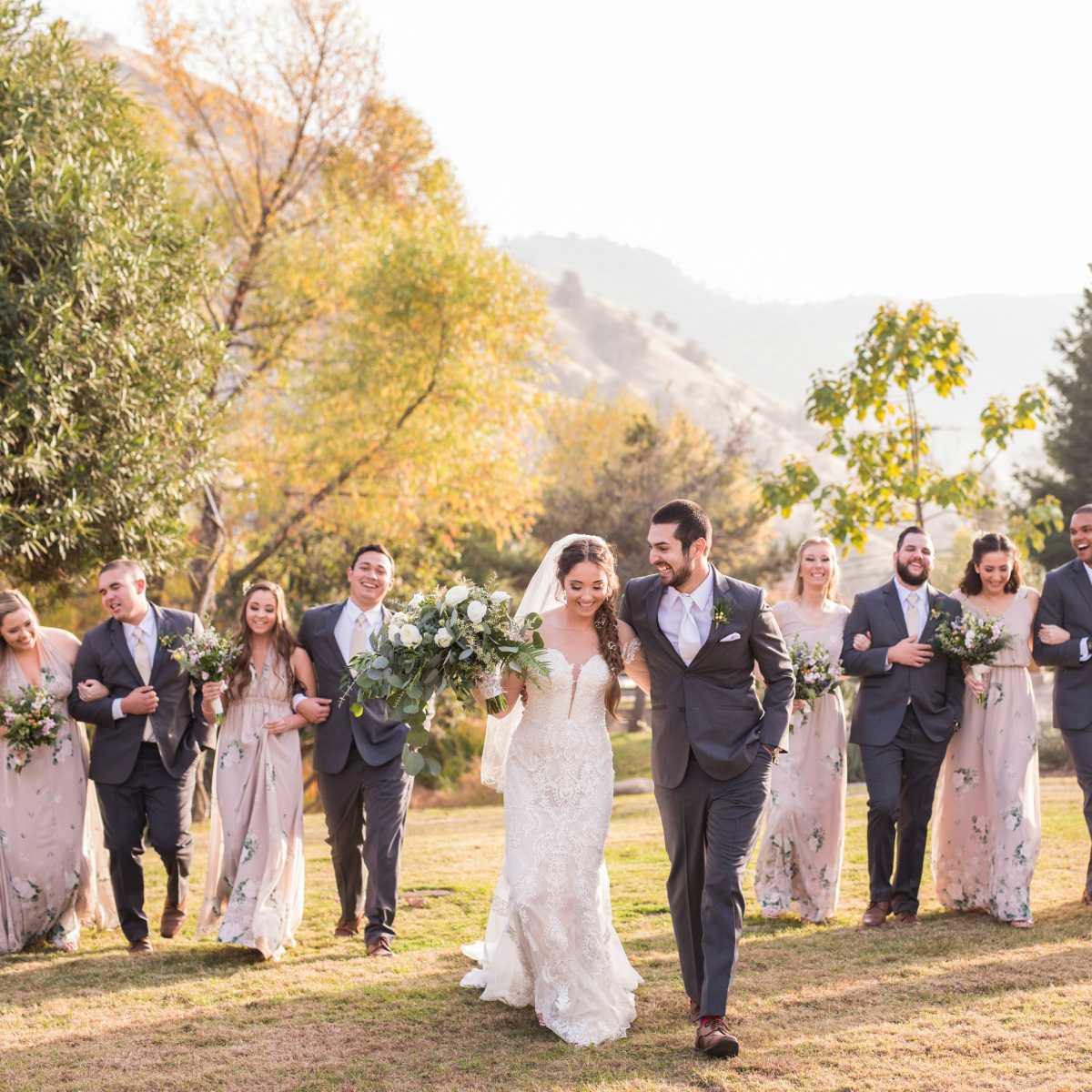 A wedding party walks together in a grassy outdoor setting, with the bride and groom leading the group.