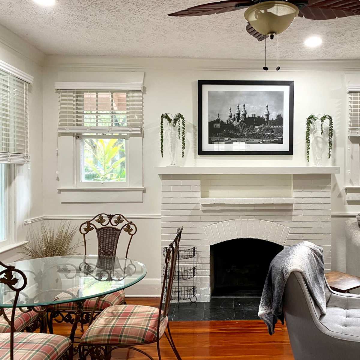 A living room featuring a white brick fireplace, a dining table with chairs, a gray sofa, and a large black-and-white photograph on the wall.