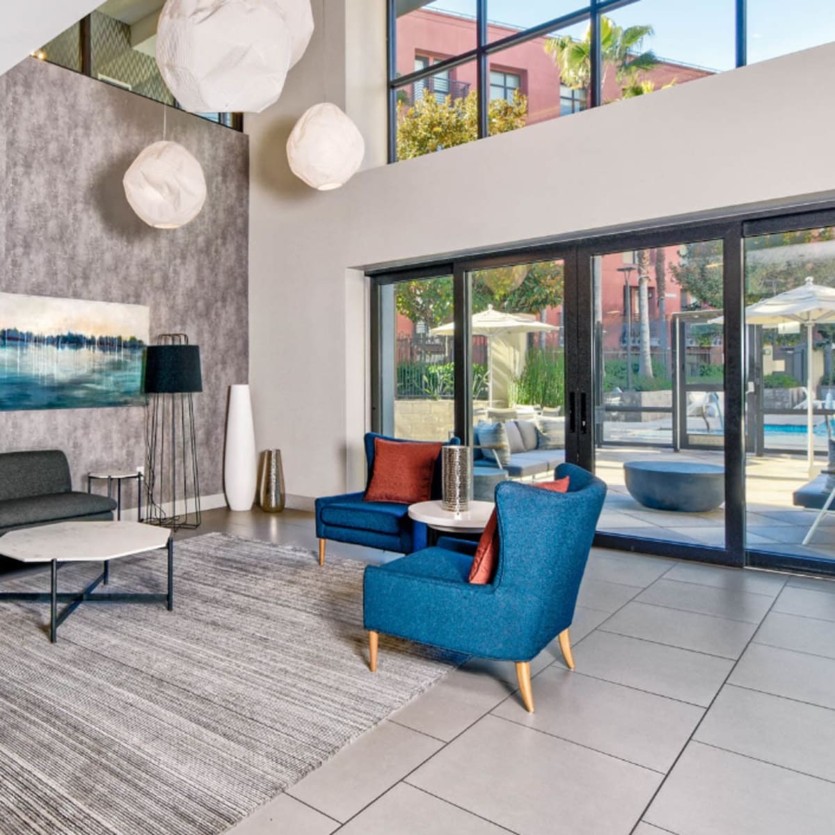 A modern lounge area features gray and blue sofas, round tables, and large windows overlooking a sunny outdoor space with patio umbrellas.