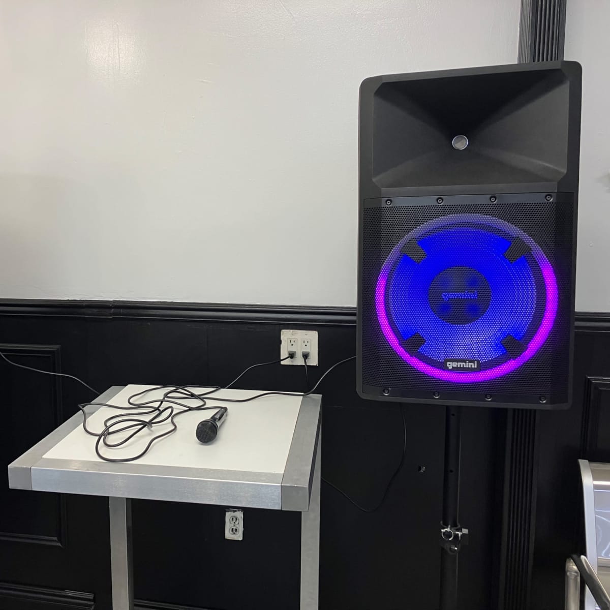 Two black speakers with colorful LED lights are positioned on stands beside a metal table in a room.