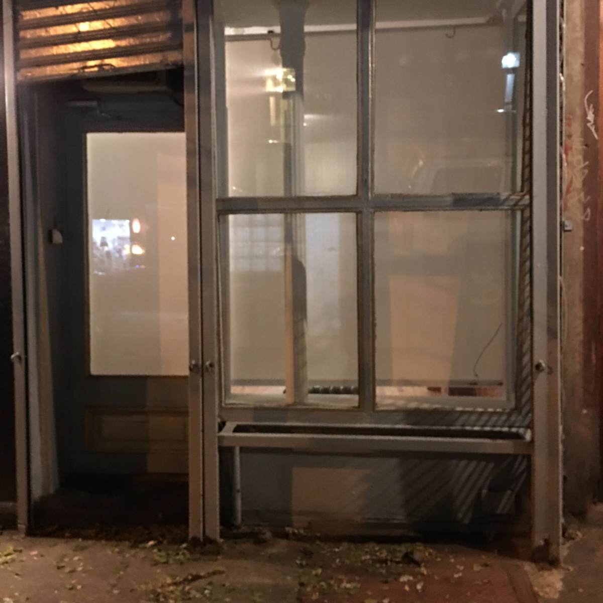 A glass-fronted storefront with a metal frame, partially illuminated at night, and surrounded by fallen leaves on the sidewalk.