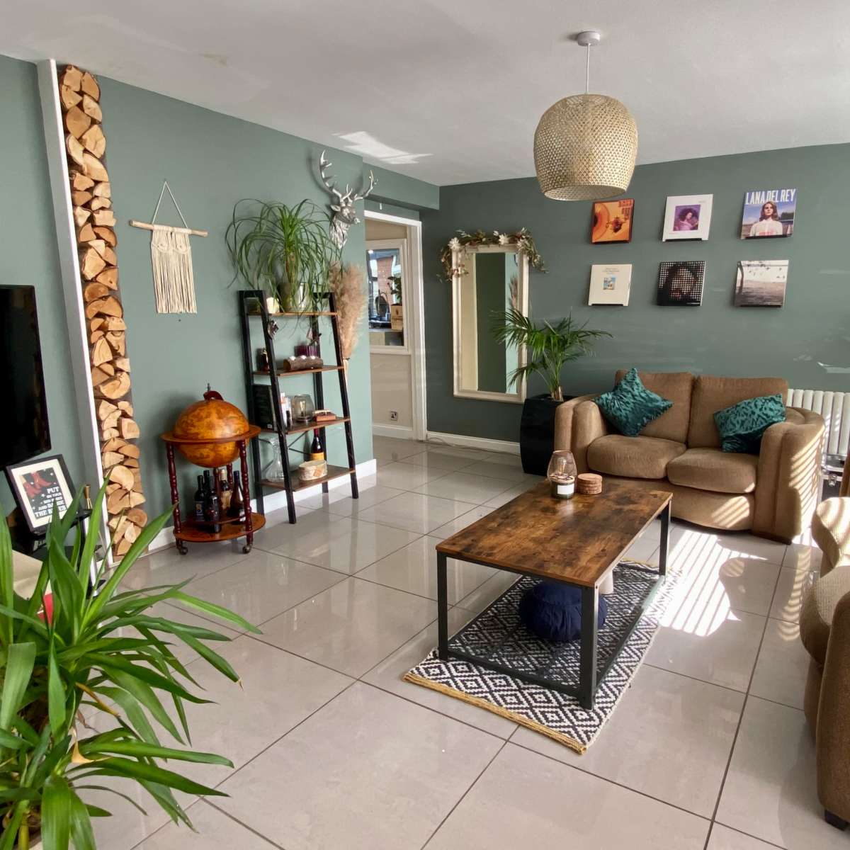 A living room with two brown sofas, a wooden coffee table, a bookshelf, and several plants, featuring light-colored tiled flooring and a green wall.