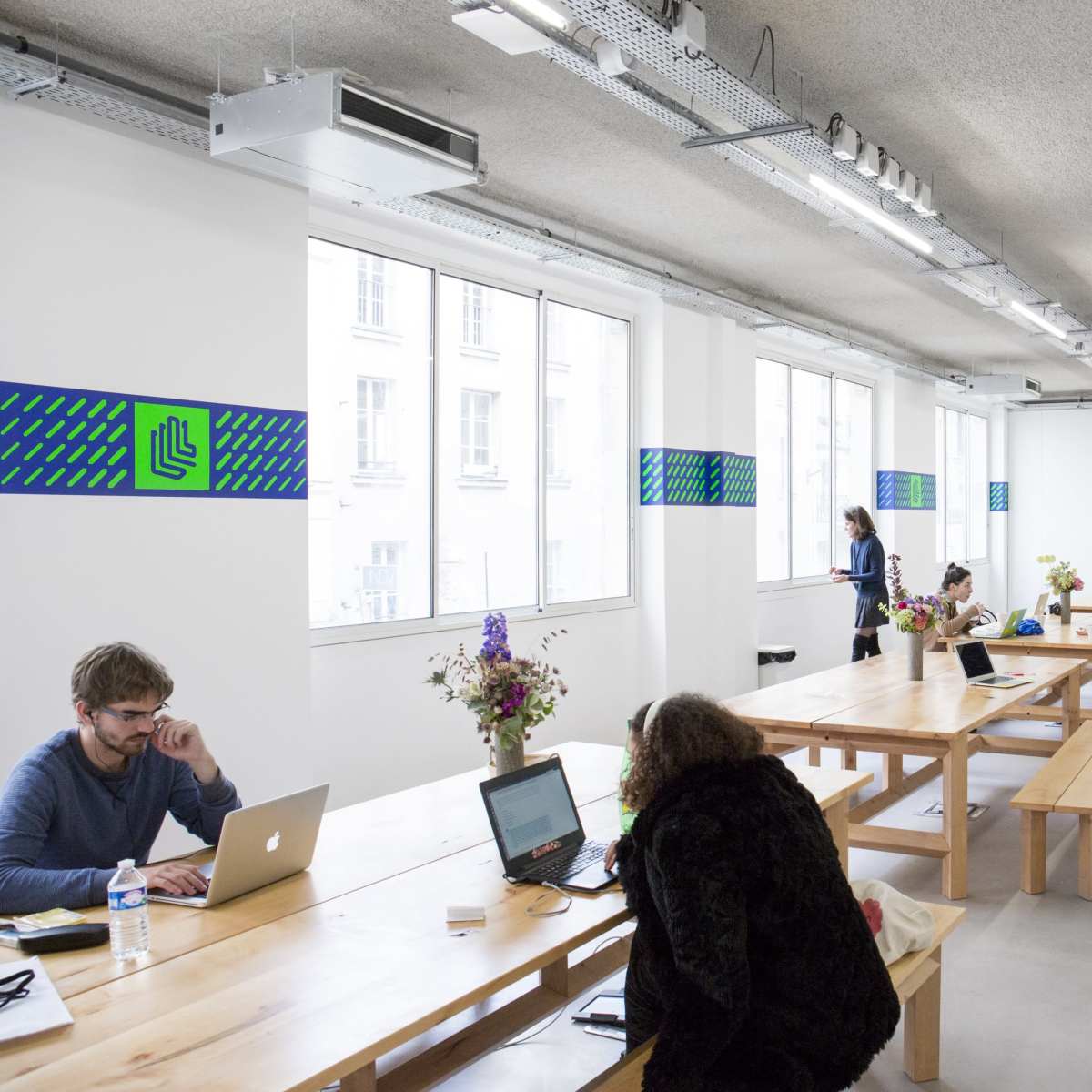 A spacious coworking space features long wooden tables, computers, and flower arrangements, with several individuals engaged in work activities.