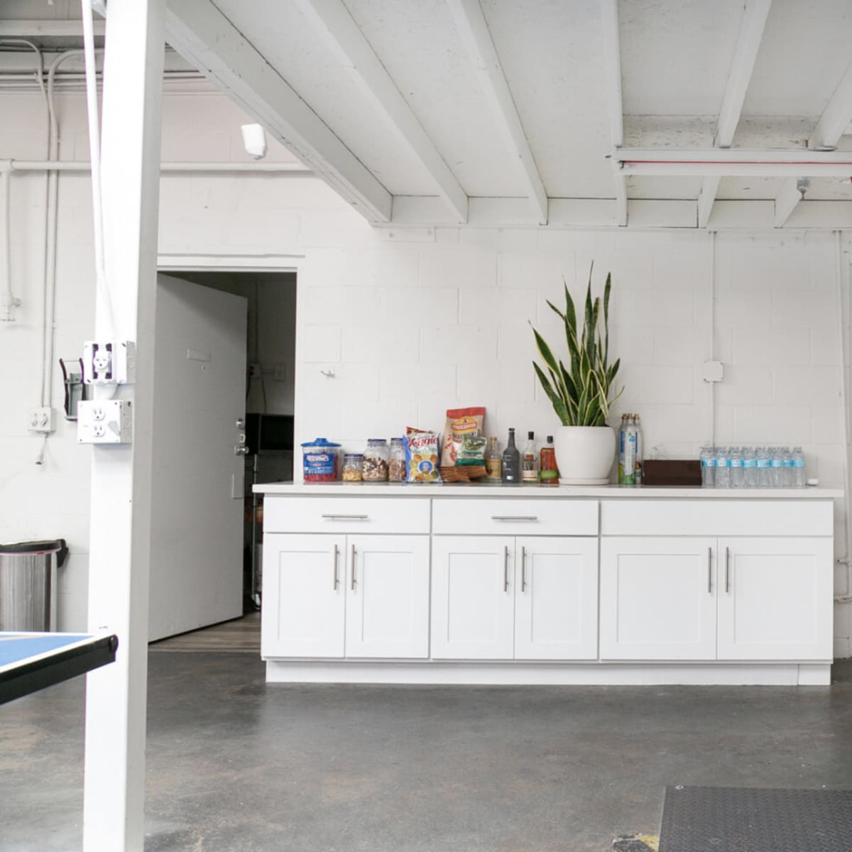 A clean garage or basement space featuring a white cabinet with snacks and water bottles, a potted plant, and an open door leading to another room.