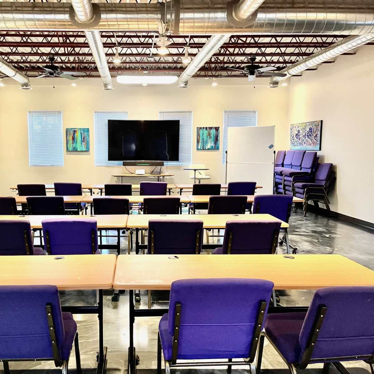 A modern classroom with rows of purple chairs and tables facing a large screen and whiteboard in a well-lit space.