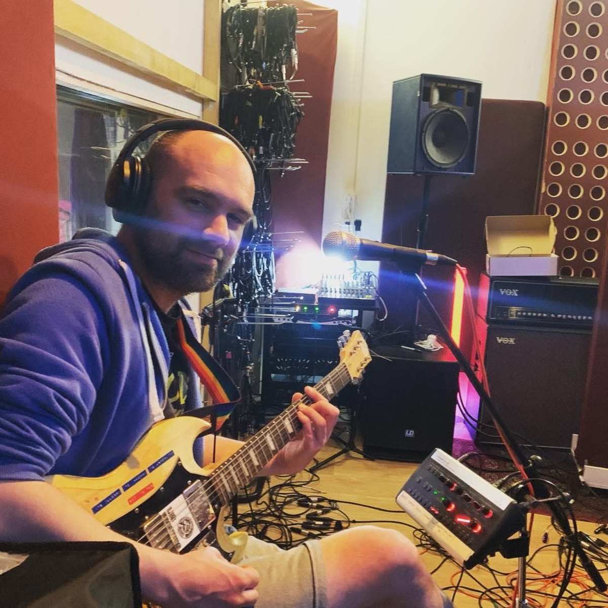 A man in a blue hoodie plays an electric guitar in a recording studio surrounded by musical equipment and cables.