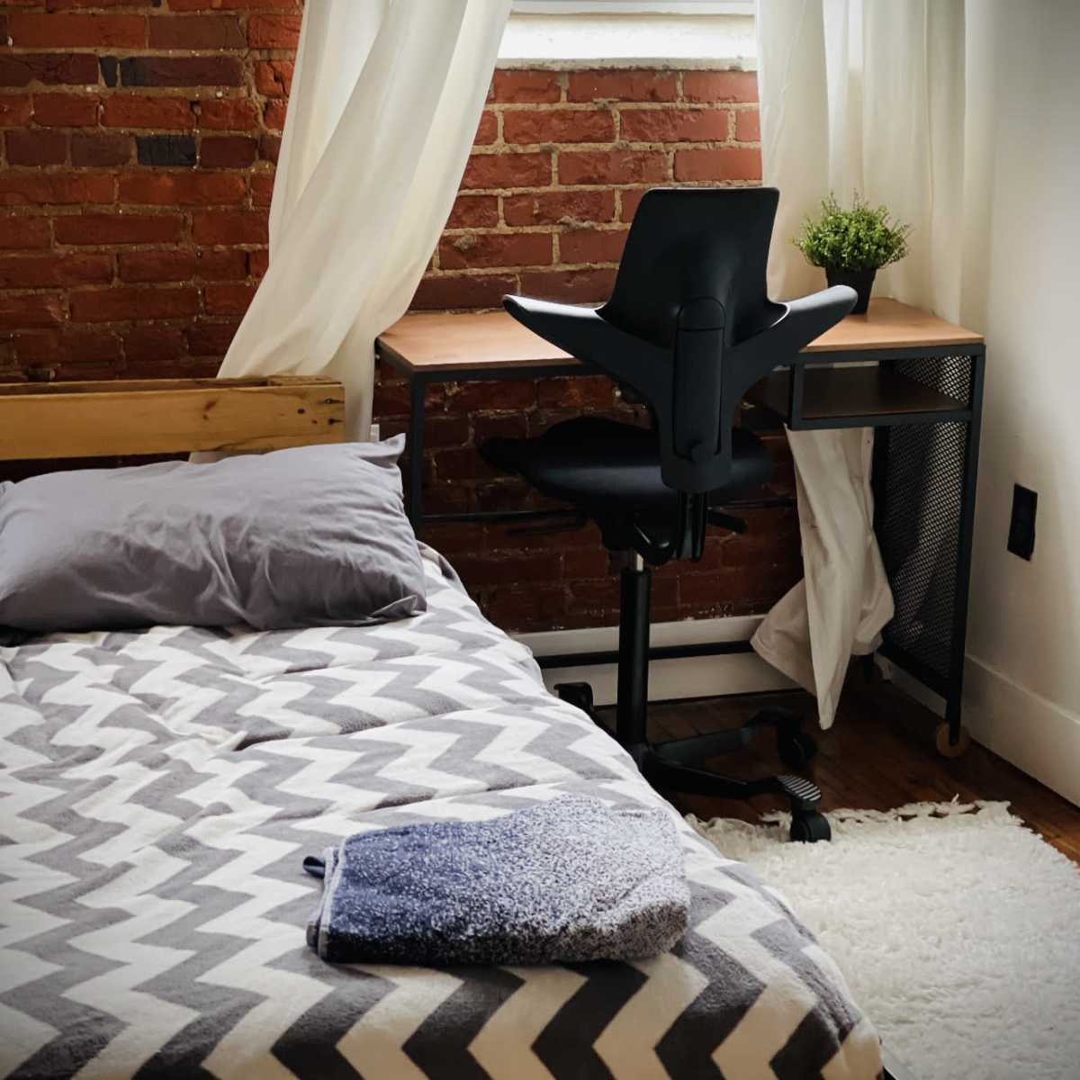 A neatly arranged bedroom features a bed with a patterned blanket and a desk with an office chair near a window draped with sheer curtains.
