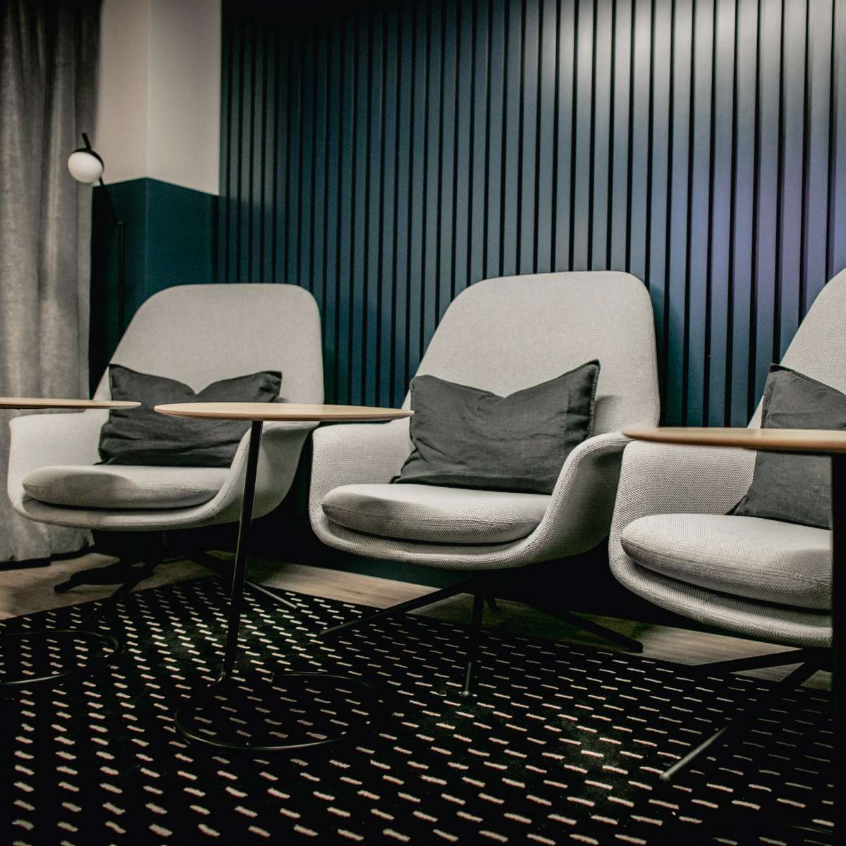 Three modern gray chairs with cushions, paired with small round wooden tables, arranged in a stylish indoor space with a textured dark wall.