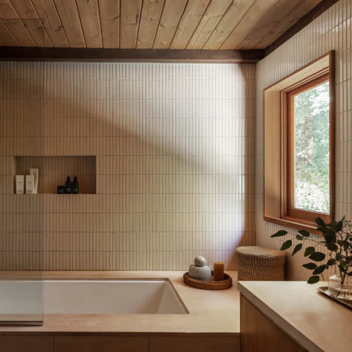 A modern bathroom with a tiled wall, a bathtub, and a wooden ceiling, featuring a window and neatly organized toiletries on a ledge.