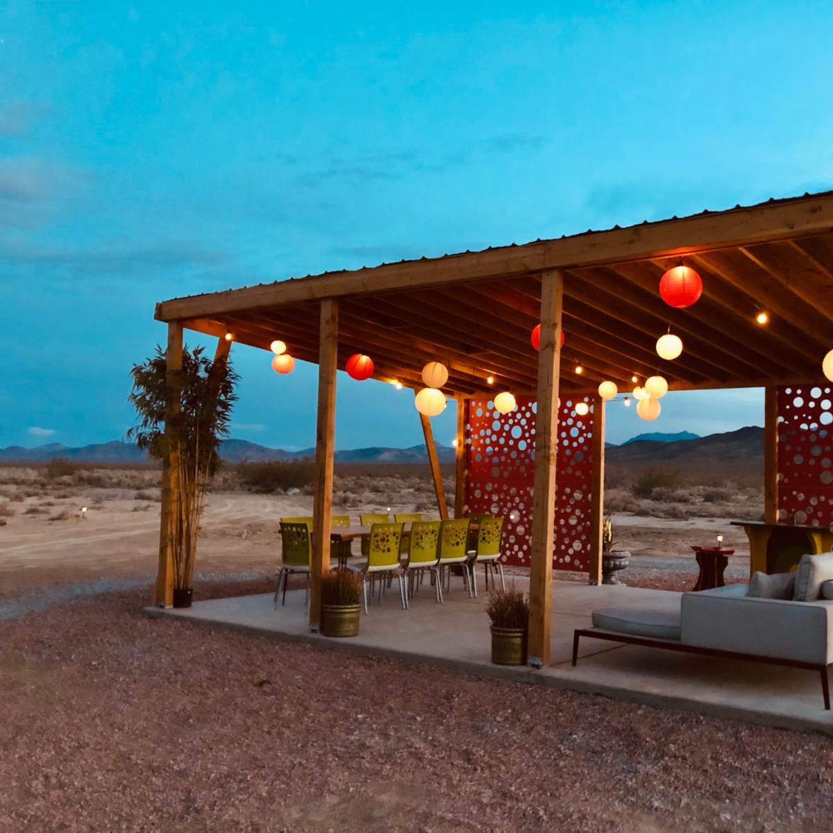 A wooden gazebo with colorful lanterns is set up in a desert landscape, featuring a seating area with a sofa and dining space for guests.