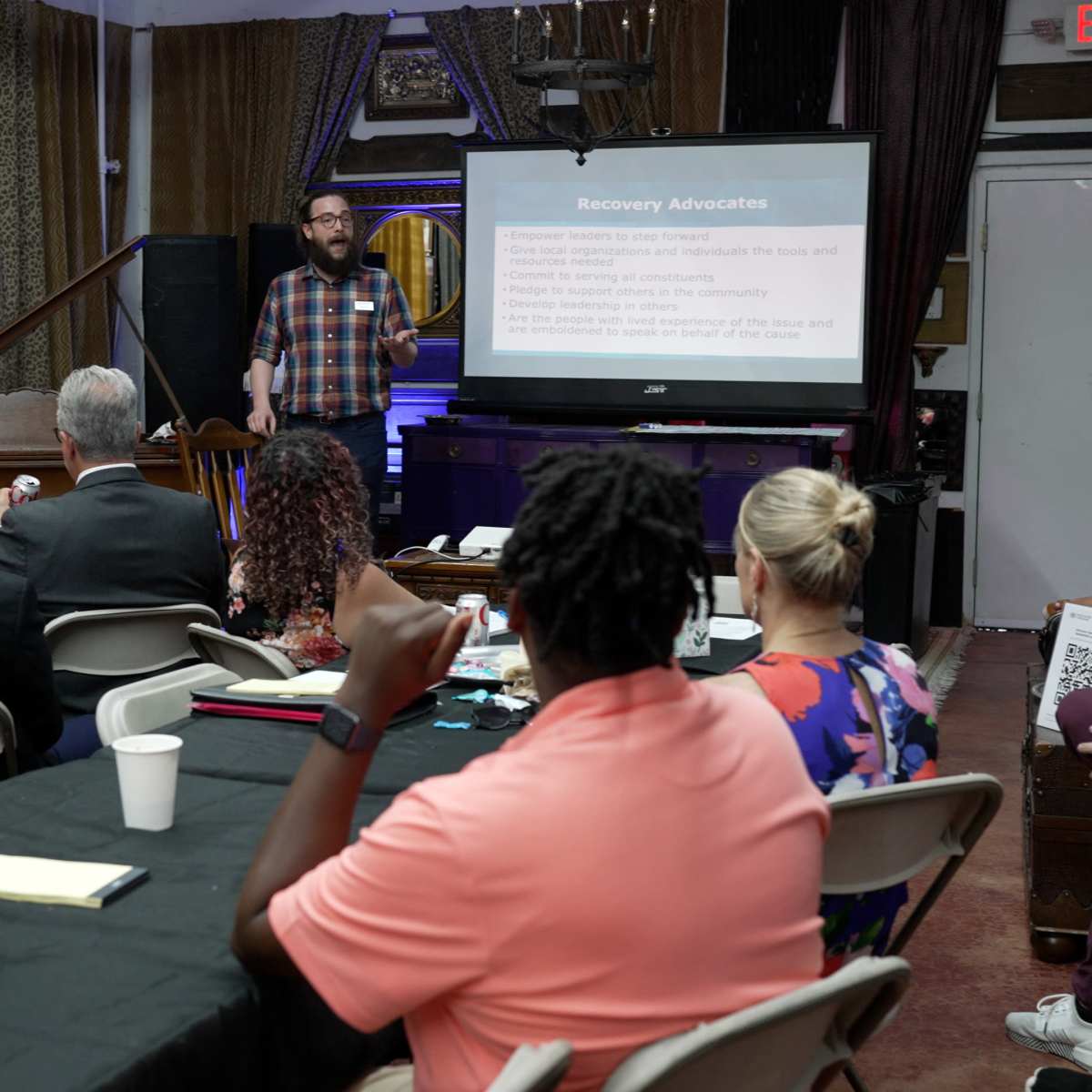 A speaker presents information about recovery advocates to an audience seated at tables in a conference setting.