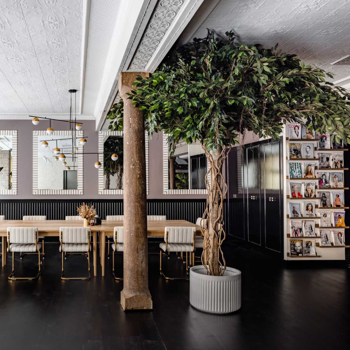 A modern dining area features a long wooden table surrounded by white chairs, with large windows and a tall potted plant enhancing the space.