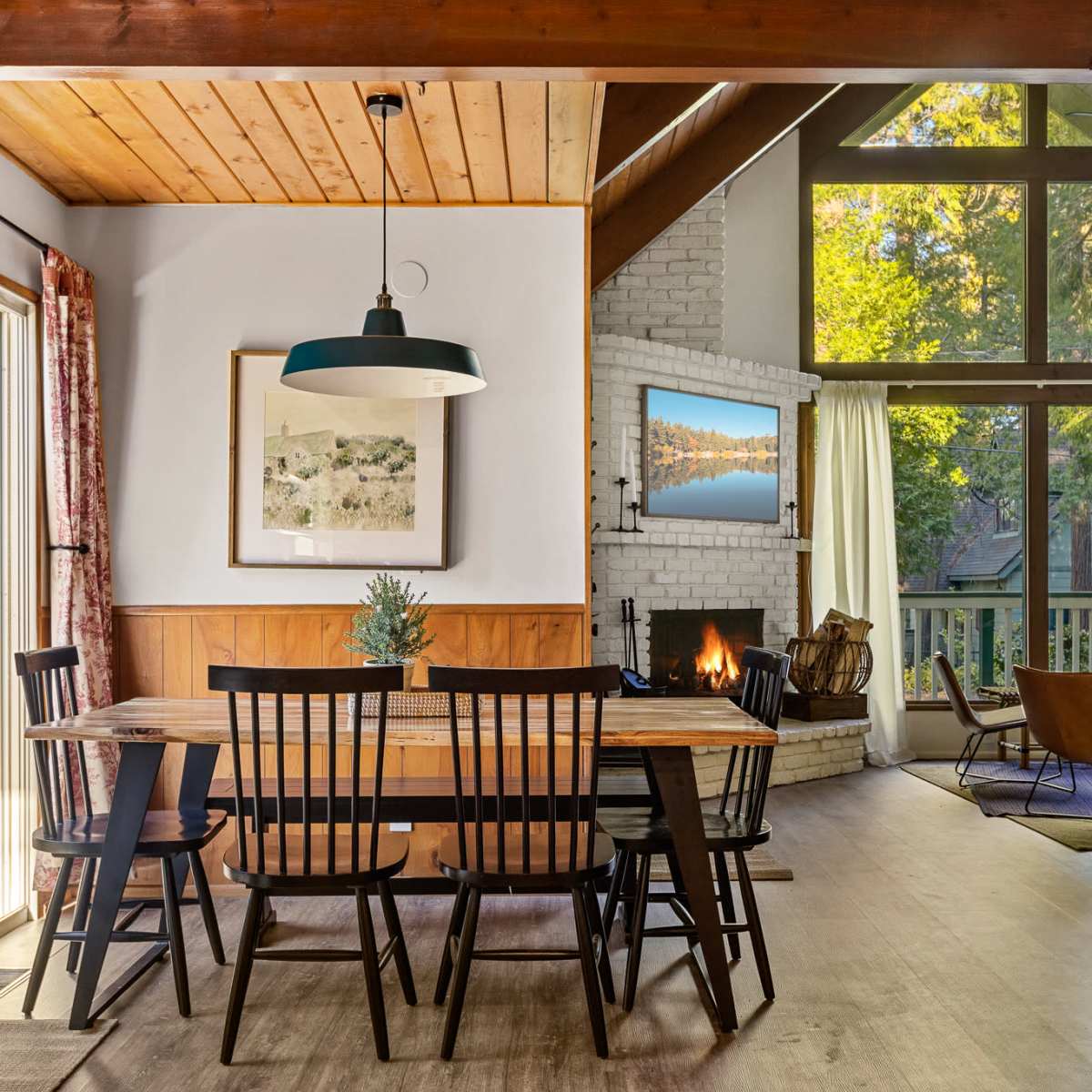 A cozy dining area with a wooden table and black chairs, adjacent to a living space featuring a fireplace and large windows that allow natural light to fill the room.