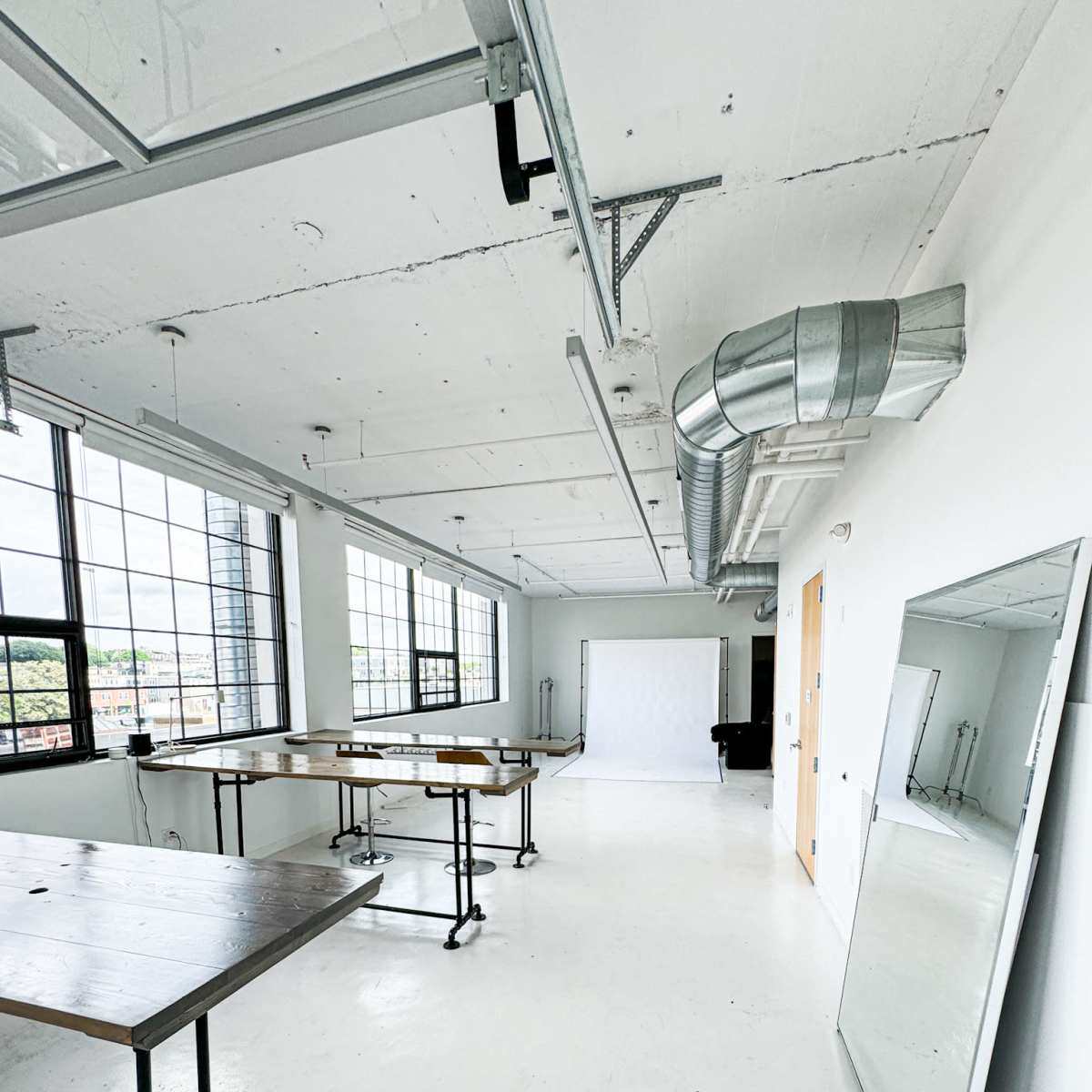 An empty studio space with large windows, wooden tables on wheels, and a white backdrop.