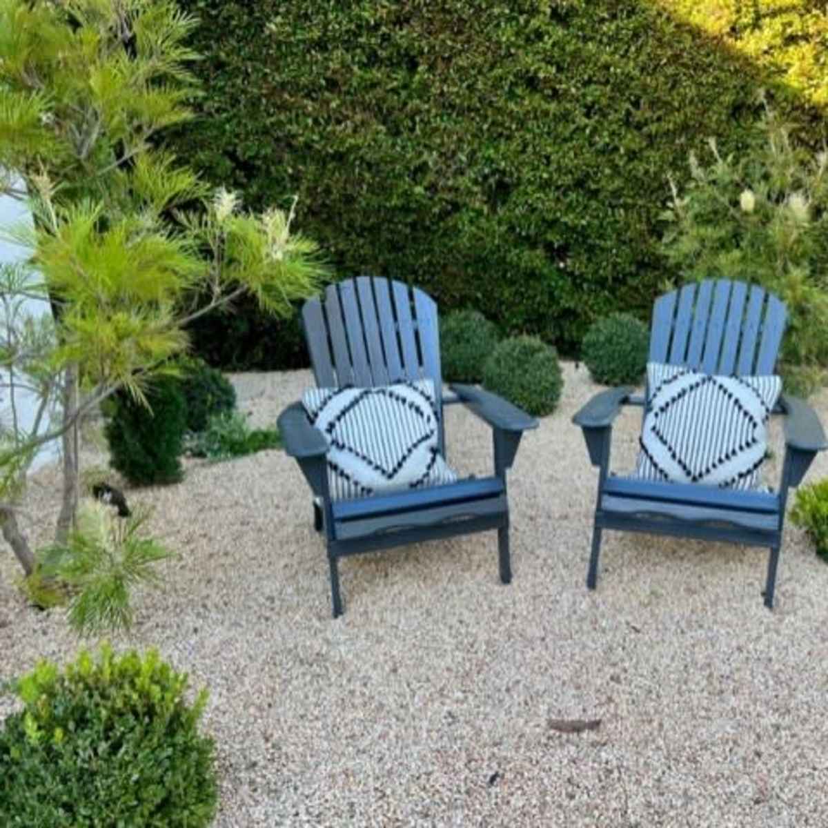 Two Adirondack chairs with patterned cushions are placed on a gravel surface surrounded by various greenery.