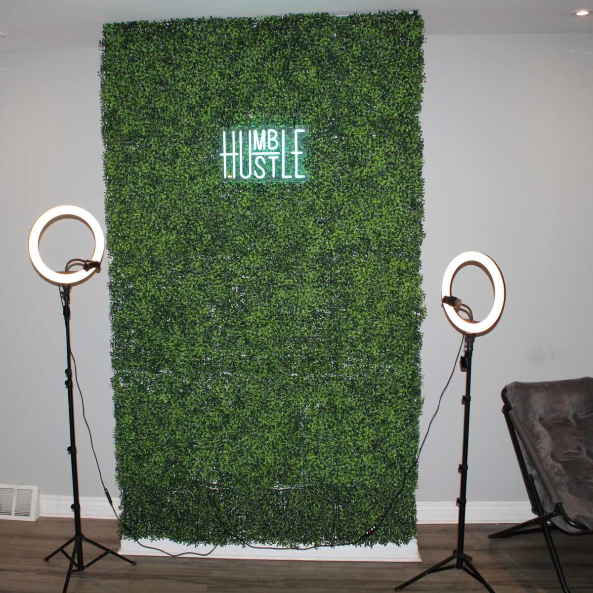 A green foliage wall with the words "HUMBLE HUSTLE" illuminated, flanked by two ring lights and a chair nearby.