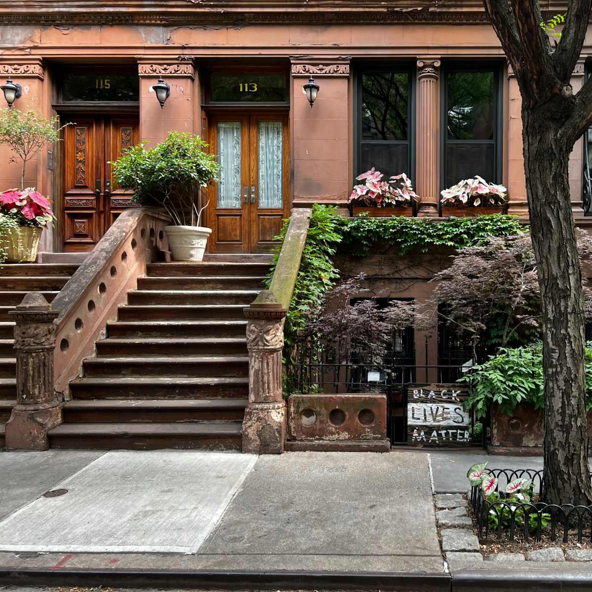 A brownstone building with a staircase, potted plants, and a "Black Lives Matter" sign in the front yard.