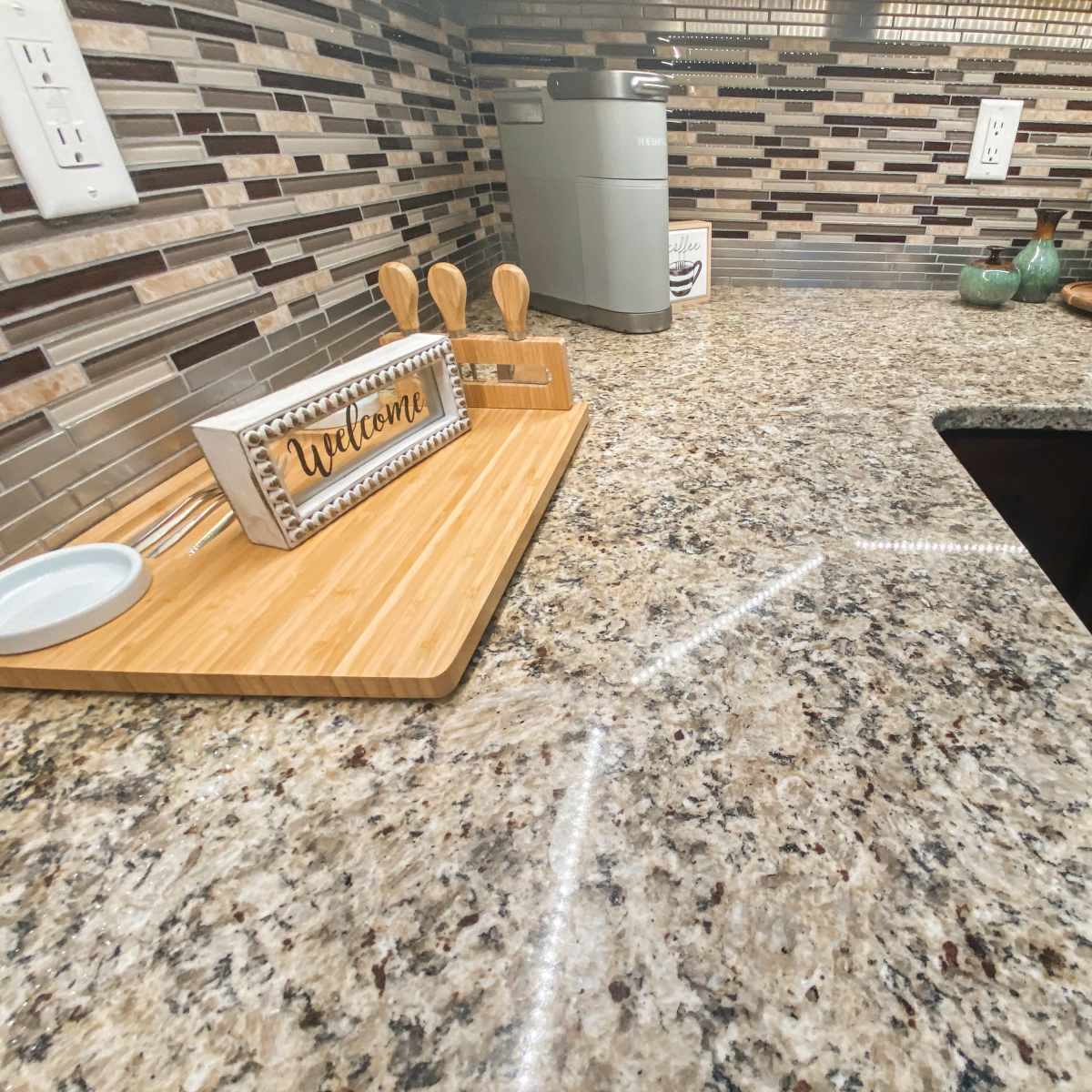 A kitchen countertop made of speckled granite, with a wooden tray that displays a "Welcome" sign and several kitchen utensils.